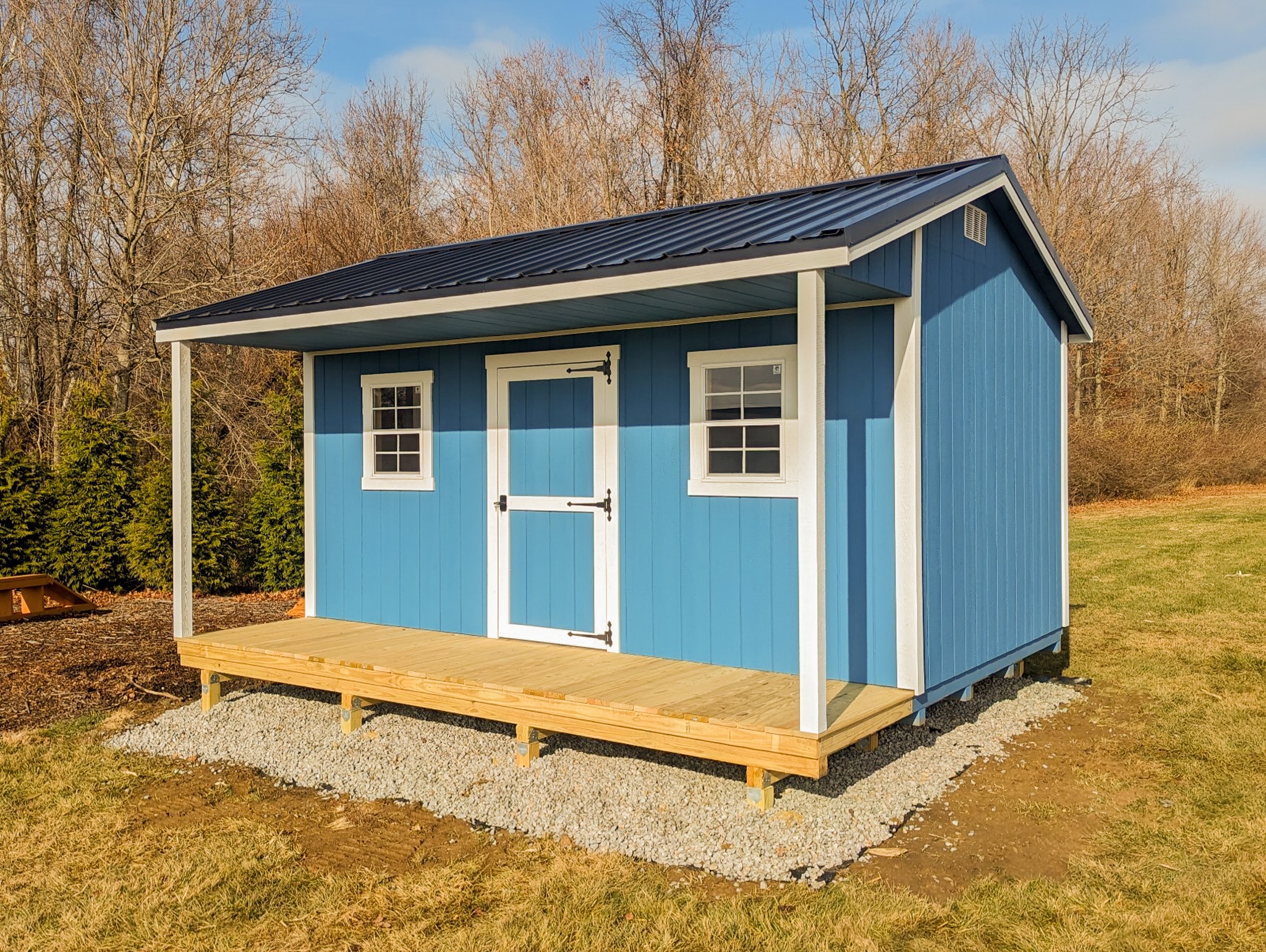 One of our blue and white cabin sheds in Gahanna OH - Blue painted wood siding with white trimmed center door flanked by two matching trimmed windows under dark metal roof - White support beams sandwiched between roof and natural wood porch