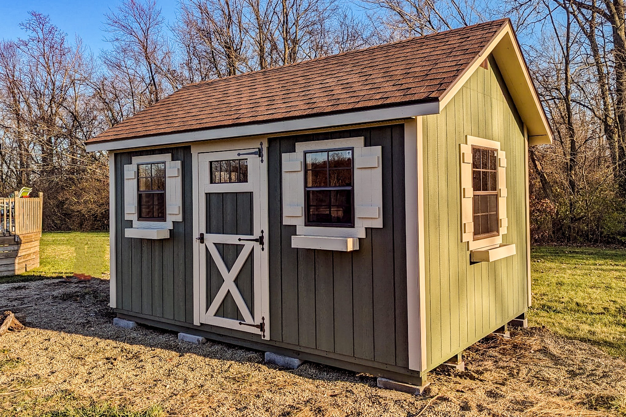 One of our green and beige Cape Cod sheds in Whitehall OH - Green siding with beige single door and three visible windows with off-white shutters beneath brown shingled roof
