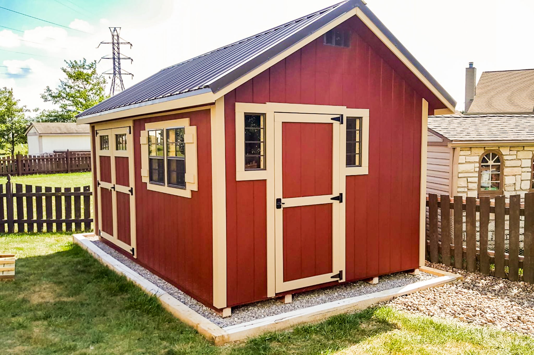 One of our Cape Cod sheds in Upper Arlington OH - Red wood siding with light peach-trimmed single and double doors - single door flanked by two small windows with peach trim - large twin windows to the right of double doors all under dark metal roof - shed sits on gravel foundation on grassy yard with brown wooden fence