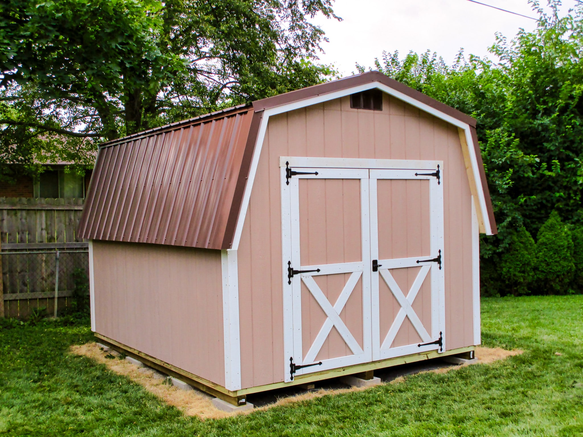 One of our classic barn-style sheds in Upper Arlington OH - peach colored wood siding with white-trimmed double doors and corners under brown metal roof - on fenced off property with green grass and verdant trees
