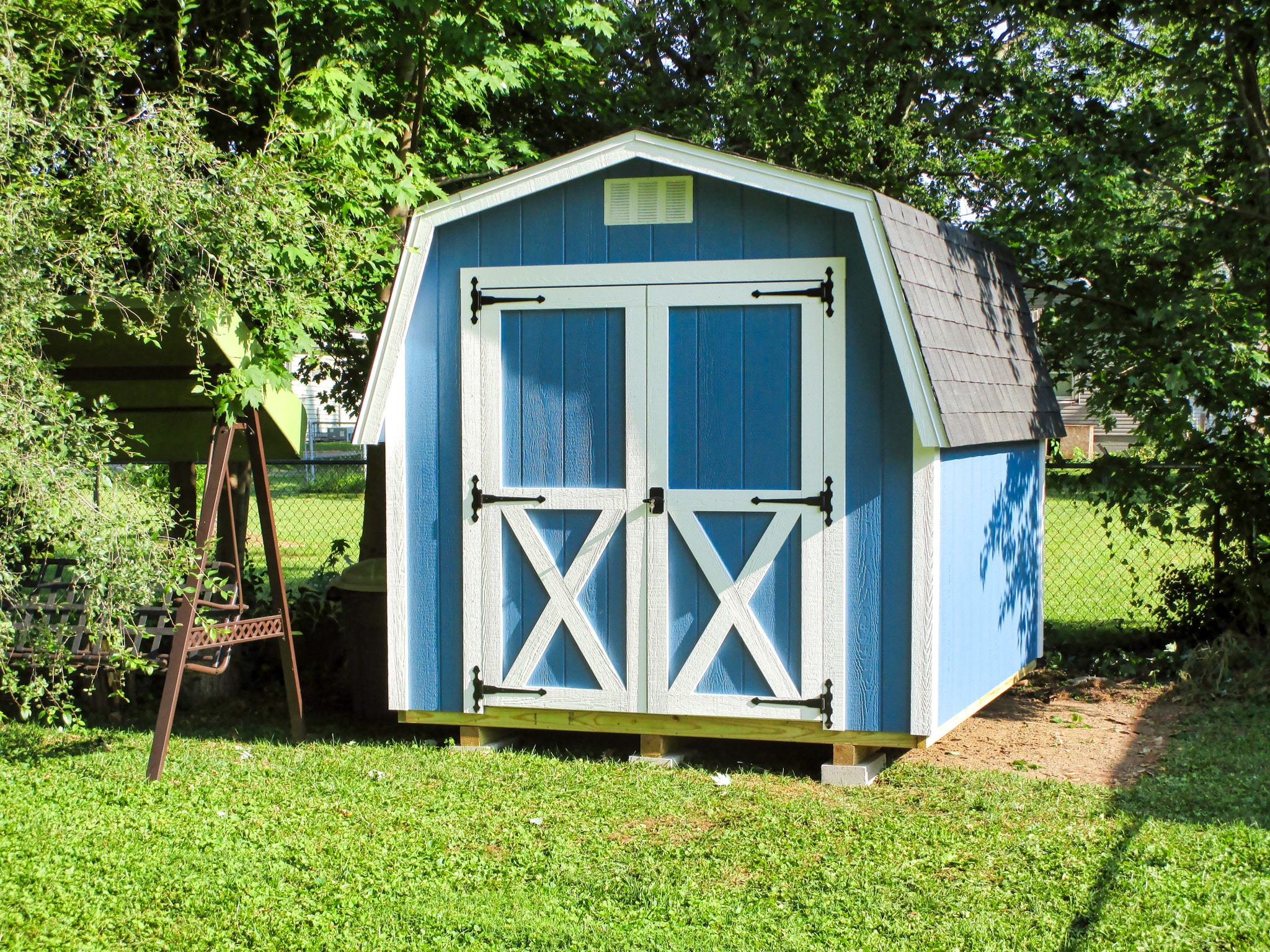 One of our Classic blue Sheds For Sale Lincoln Village OH - Blue siding with white trimmed double doors under white vent and gray roof - set on green grass amid trees