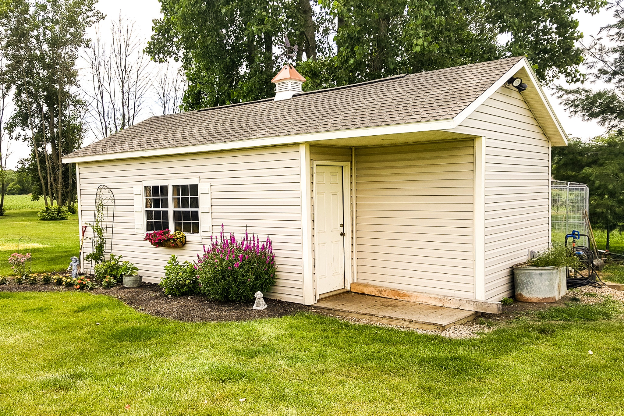 One of our large custom sheds in Whitehall OH - Eggshell siding with white-shutter windows with matching door and corner trim. Window has flowers in a windowbox - shed is topped with brown shingled roof with a white and copper colored cupola - shed sits on grassy property with nearby trees