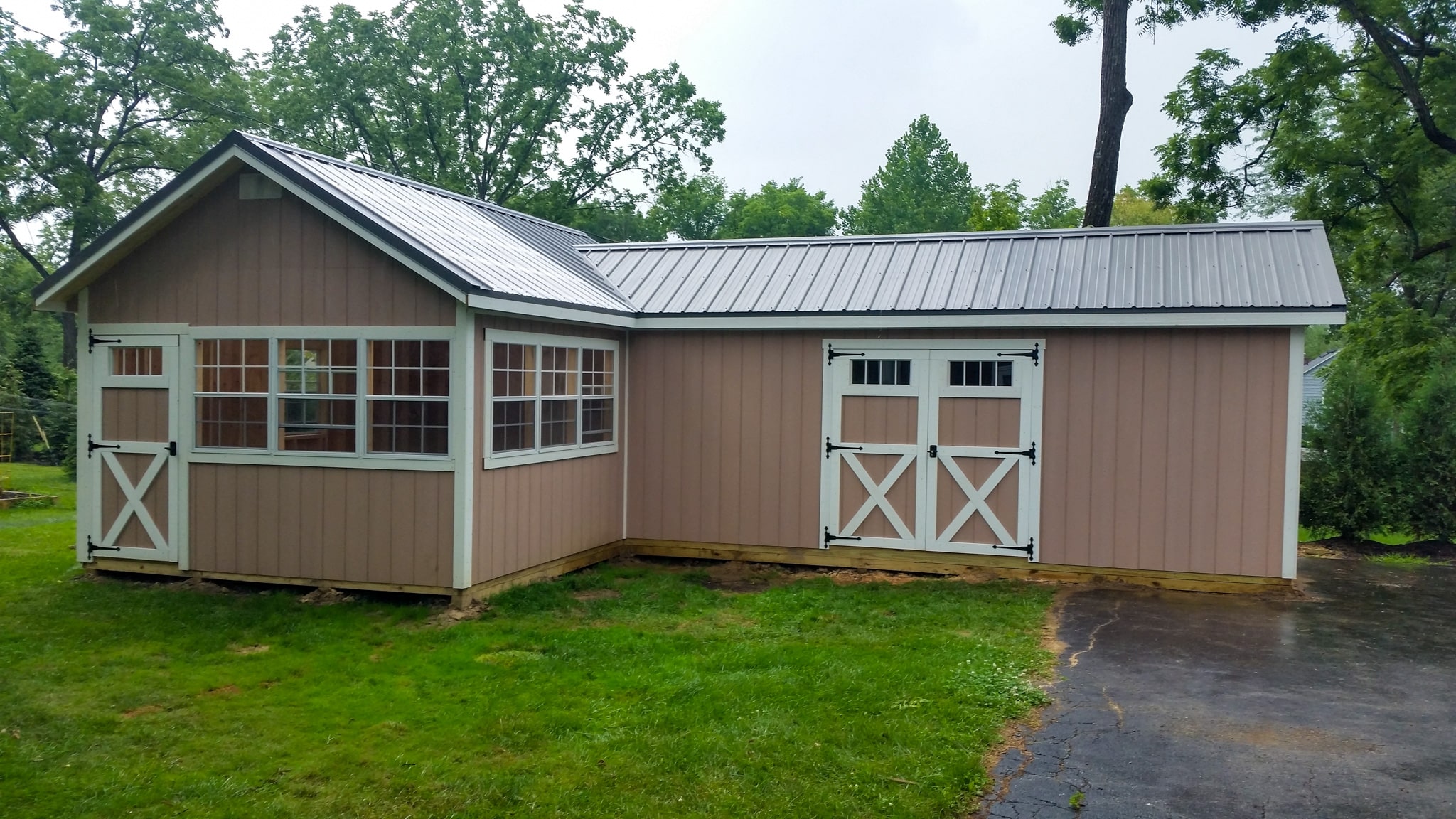 One of our peach and white custom sheds for sale in Lincoln Village OH - Peach L-shaped shed with white-framed double doors on longer side single white-framed windows and door on shorter side under dark gray roof - set on green grass and dark pavement amid trees on cloudy day