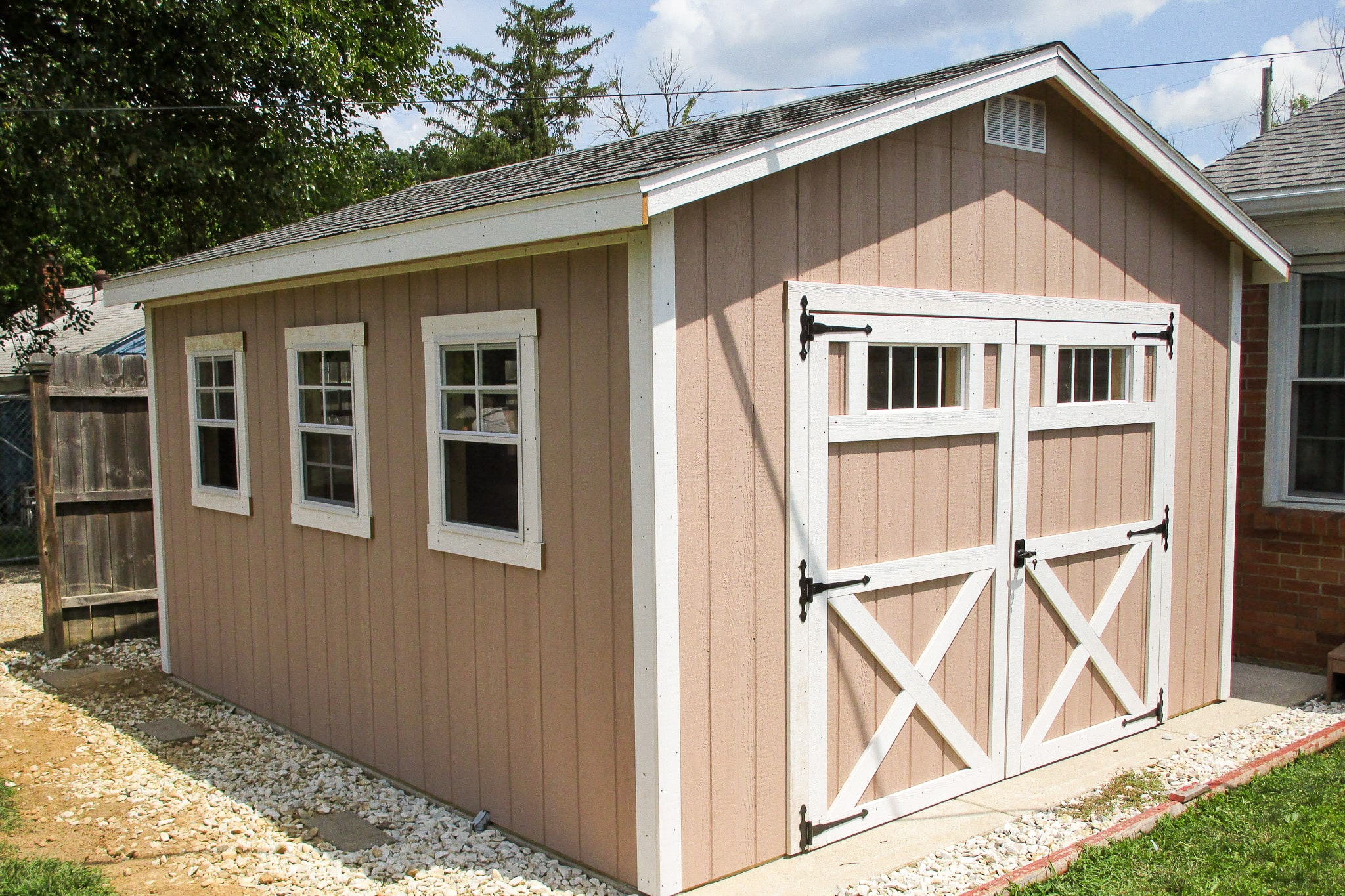 One of our peach and white gable sheds in Gahanna OH - Peach siding with white-trimmed double doors with X detail and three matching windows - gray shingled roof - shed sits on concrete bordered with green grass with trees in the background