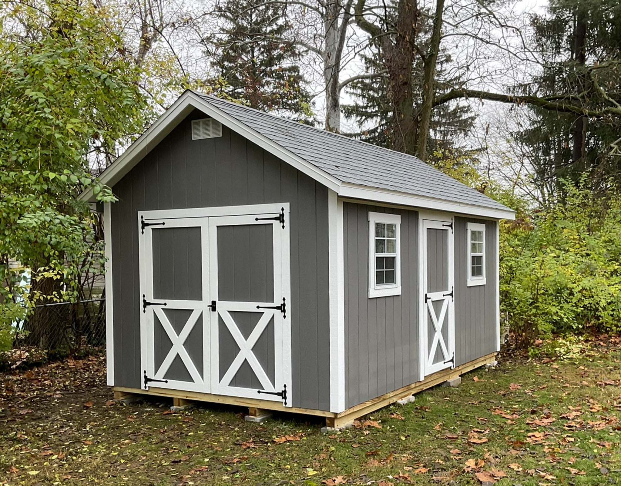 One of our gray and white gable sheds - gray siding with white-trimmed double and single doors and windows beneath gray roof - set on grassy area lined with trees