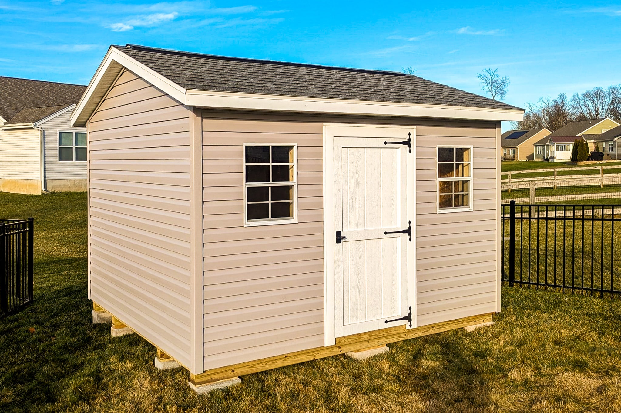 One of our beige gable sheds in Whitehall OH - beige siding with white door between two white-framed windows beneath dark gray shingled roof - on black-gated lawn with houses in the distance