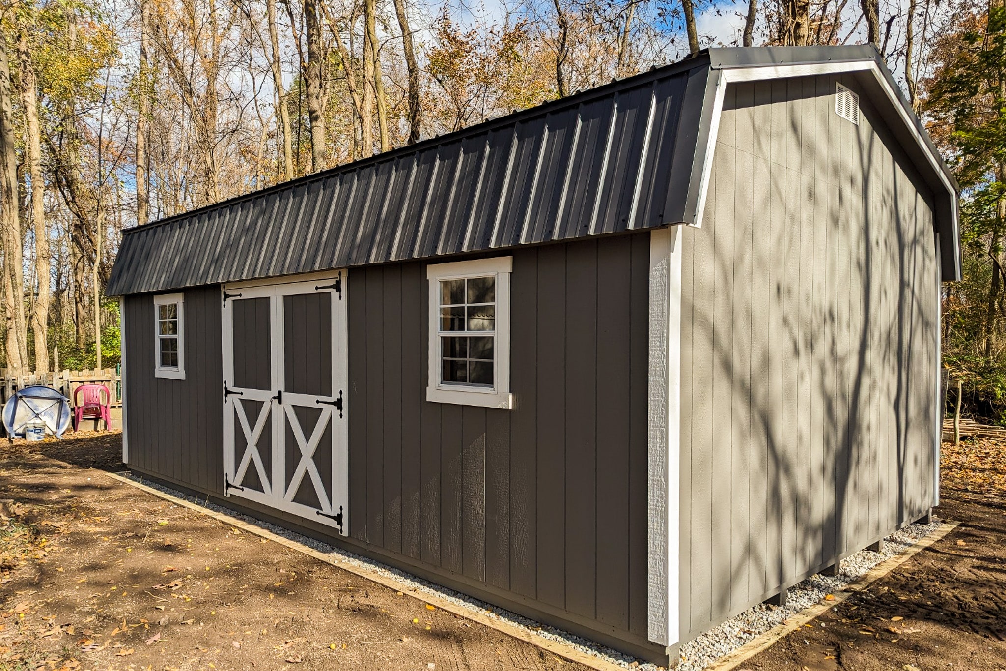 One of our dark gray highwall sheds in Gahanna OH - Dark gray wood siding with white-trimmed double doors between two white-framed windows beneath gray metal roof - shed sits on gravel foundation surrounded by brown earth on tree-lined property