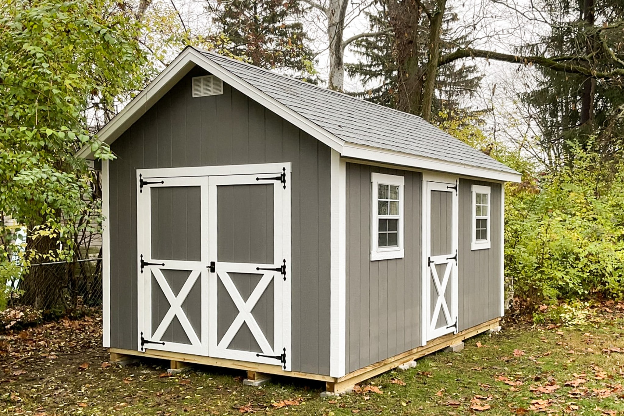 One of our gray and white Cape Cod Sheds in Gahanna OH - Gray wood siding with white-trimmed single and double doors and white trimmed windows under gray shingled roof - shed sits on grassy corner of yard lined with trees