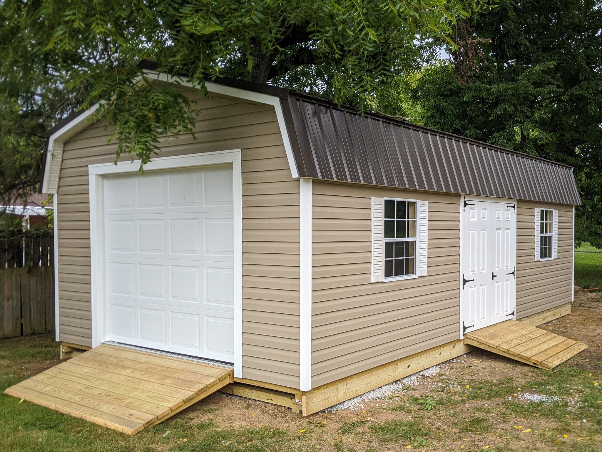 One of our beige and white highwall garage sheds - beige siding with white double doors between two windows with white shutters - white overhead door beneath brown metal gambrel roof - shed sits beneath canopy of green leaves