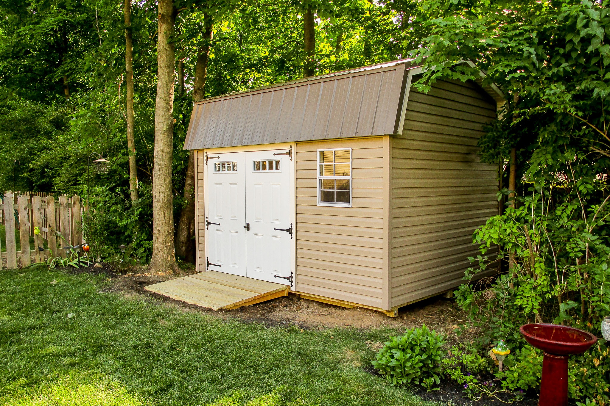 One of our highwall sheds in Whitehall OH - beige siding with white glass-inset double doors with black hinges - single white-framed window all beneath brown metal gambrel-style roof - shed is nestled among greenery in backyard - red bird bath in lower right portion of image