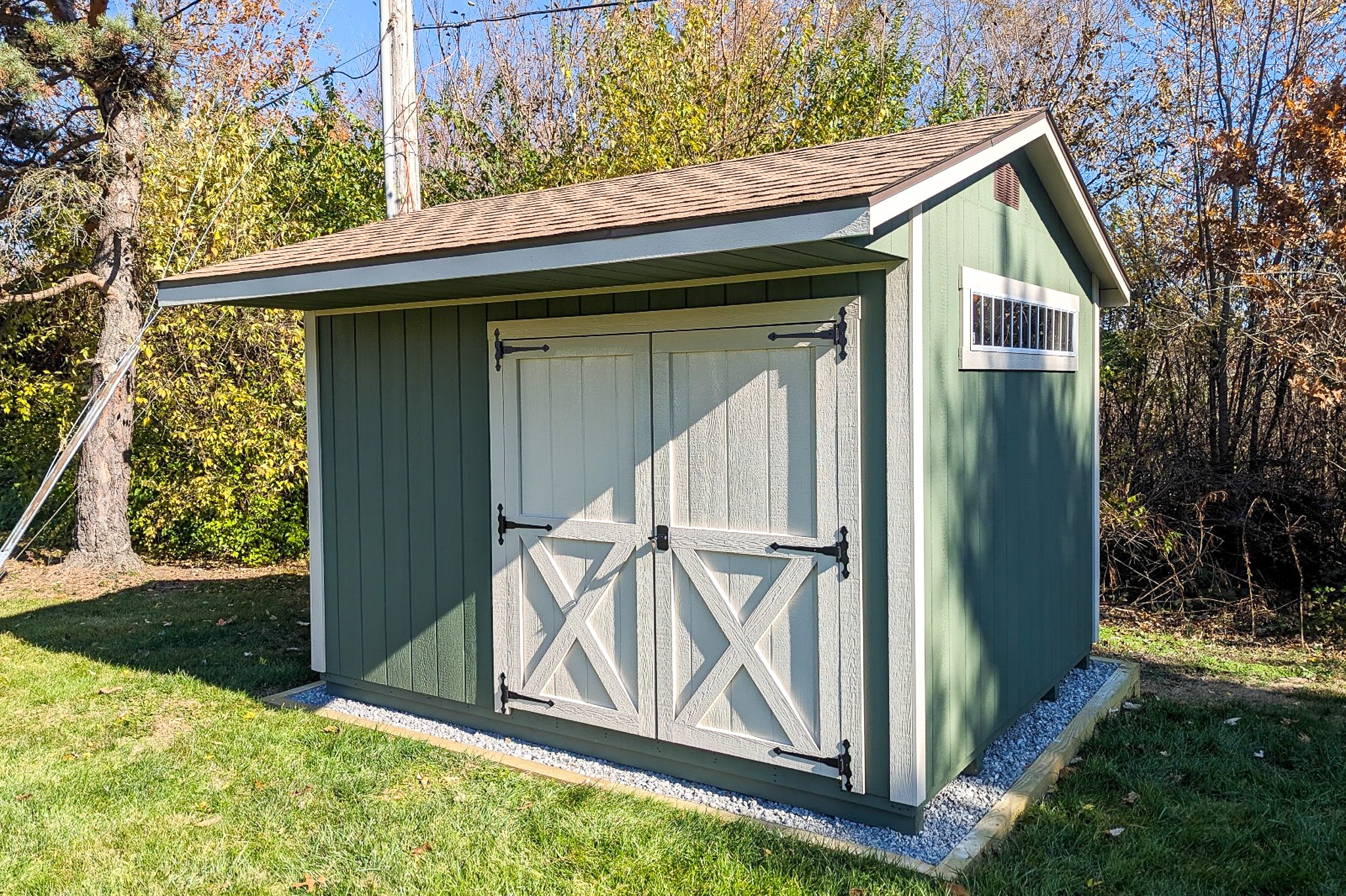 One of our green and white wood Quaker sheds in Whitehall OH - medium green siding with white double doors and corner trim beneath brown shingled overhanging roof - horizontal row of glass panes on right external wall