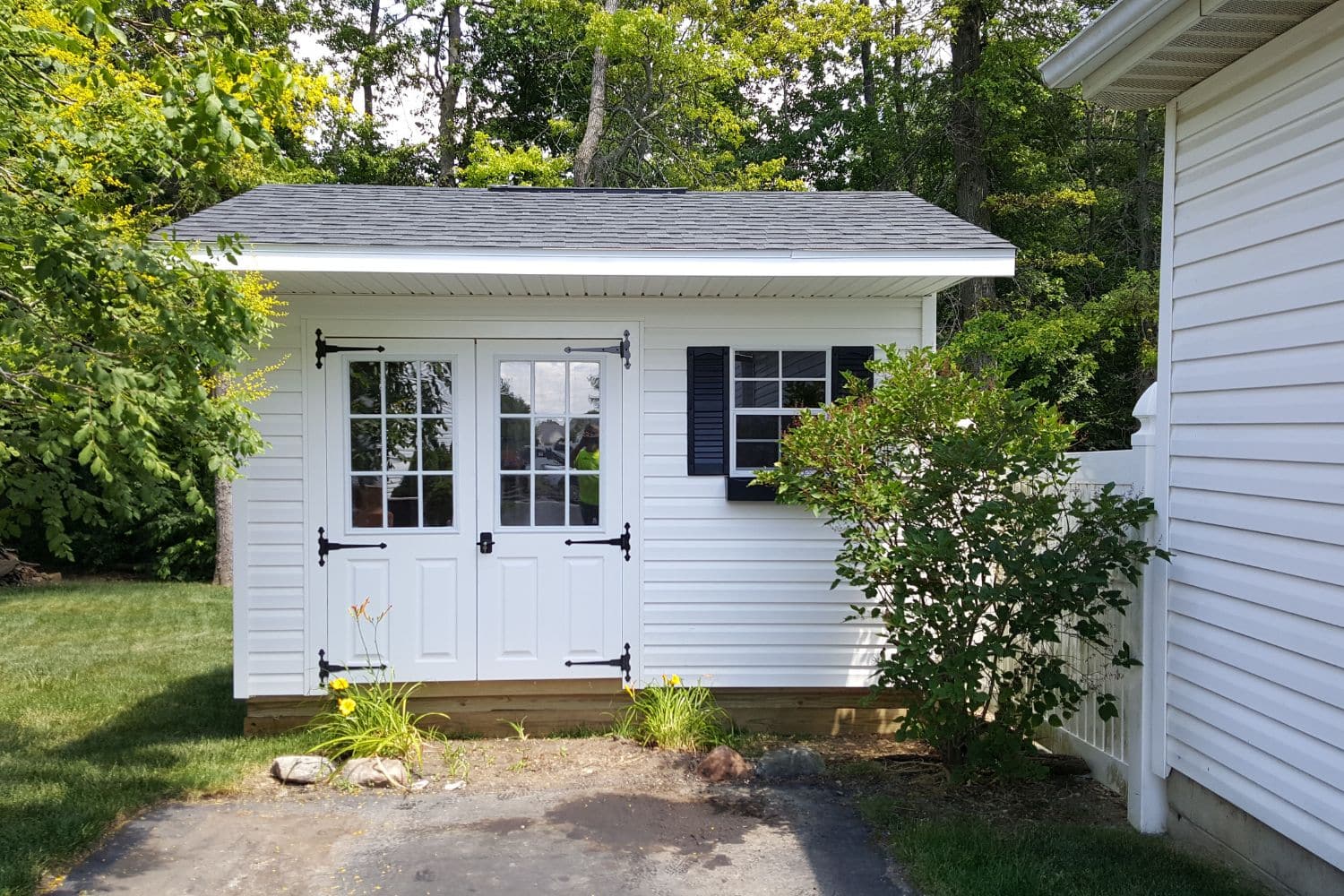 One of our white Quaker sheds for sale in Worthington OH - White siding with white trimmed window and double doors - gray roof - shed is nestled among green trees