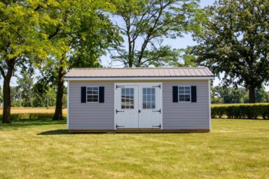 One of our gable sheds for sale in Dublin OH - Beige siding with white doors white trimmed windows and black shutters - brown sun-lit roof - green lawn hedges and trees