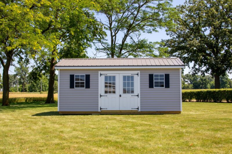 One of our gable sheds for sale in Dublin OH - Beige siding with white doors white trimmed windows and black shutters - brown sun-lit roof - green lawn hedges and trees