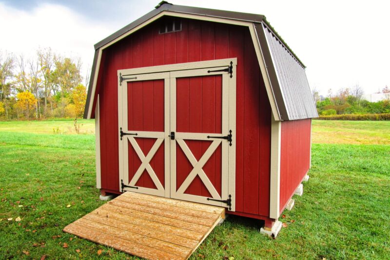 One of our classic barn sheds for sale in Dublin OH - Red siding with beige double doors behind a wooden ramp - brown rood - bright green grass on mostly cloudy day