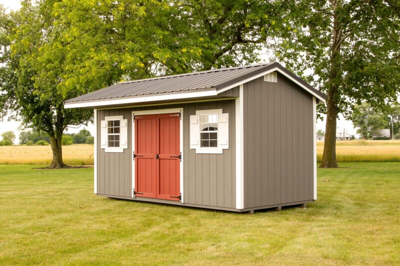 A gray Quaker shed with a distinctive red double door and white trim, featuring round accent windows, commonly seen in our Quaker sheds in Blacklick Estates, situated on a neatly mowed lawn surrounded by fields and mature trees.