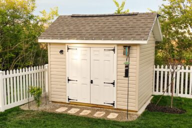 A beige shed with white double doors and elegant lantern-style lights, typical of our quality sheds in Blacklick Estates, set on a stone walkway surrounded by a white fence and lush greenery.