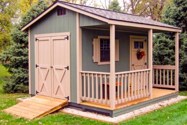 A charming, cabin-style shed with a green exterior and beige trim, featuring a small front porch with railings and double doors, typical of our popular sheds in Blacklick Estates. The shed is set on a wooden ramp and surrounded by neatly cut grass and trees in a peaceful setting.