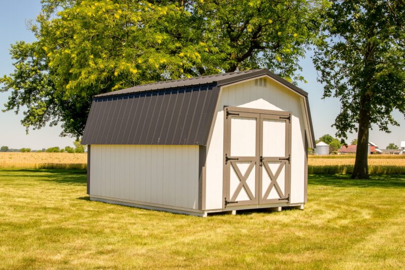 A small, white barn-style shed, typical of sheds in Blacklick Estates, with double doors and a dark metal roof, standing on a well-manicured lawn surrounded by a few large trees. In the background, there is a field of crops and some farm buildings under a clear blue sky.