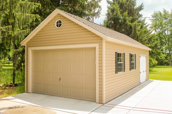 A tan Gable-style garage shed with a large roll-up door, black-framed windows, and a small circular vent at the peak, typical of our garages in Blacklick Estates, set on a concrete driveway surrounded by trees.